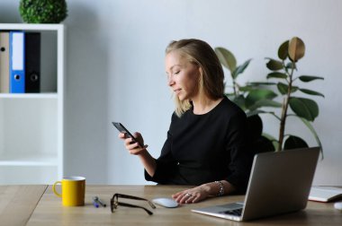 Masasındaki profesyonel kadın, modern ofis ortamında dizüstü bilgisayarı, bardakları ve kahve bardağıyla akıllı telefonunu kontrol ediyor..