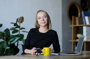 Modern bir ofiste elinde kahve fincanı, bardak ve bilgisayarla gülümseyen ve akıllı telefonu tutan profesyonel bir kadın..