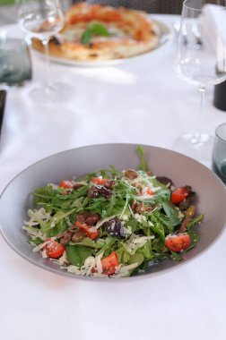 Beyaz masa örtüsünde servis edilen peynirli ve zeytinli taze bir salata ve arka planda pizza..