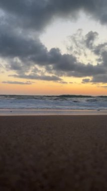Gün batımında kumlu bir sahile vuran okyanus dalgalarının alçak açılı görüntüsü. Gökyüzünde dramatik bulutlar var..