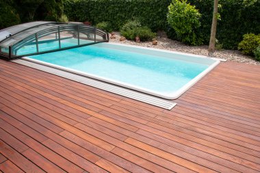 Outdoor swimming pool with cover enclosure and teak wood deck, exotic teakwood flooring stripes around blue water pool clipart