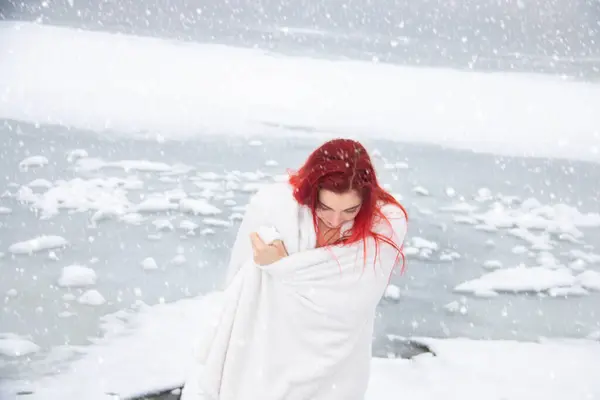 Winter swimming and cold water immersion. Red hair girl wrapped in a towel is hardening outdoors on snow at white winter lake landscape, while snow is falling