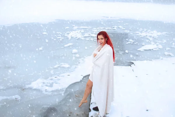 Ice woman wrapped in a towel dipping a foot into cold water while snowing outdoors at frosty lake, cryotherapy and winter swimming lifestyle
