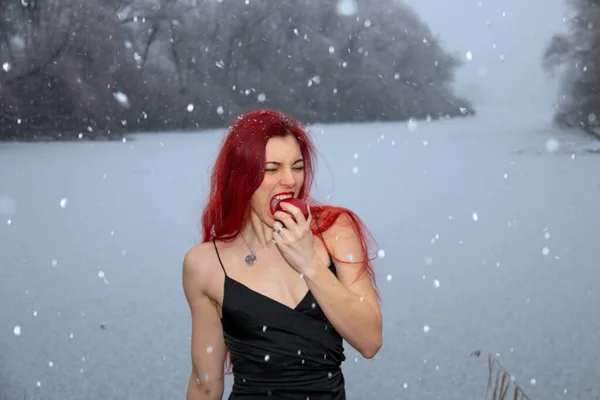 Woman in a black dress is eating red apple outdoors near frozen lake winter background while snowing