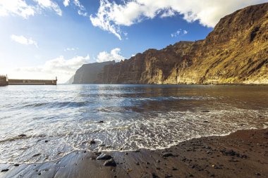 Dik sarı uçurumlar ve limana bakan manzaralı koyu kumlu ve kayalık plajlar. Fotoğraf 4 Haziran 2022 'de Tenerife, İspanya' daki Los Guios plajında çekildi..