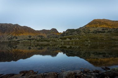 Güneşin yumuşak, turuncu ışıkla doğuşu Retezat dağlarının tepelerini aydınlatıyor ve Bucura Gölü 'ne yansıyor. Fotoğraf 7 Ağustos 2022 'de Romanya' nın Retezat kentinde Bucura Gölü kıyısında çekildi..