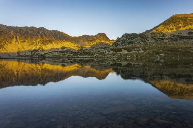 Güneşin yumuşak, turuncu ışıkla doğuşu Retezat dağlarının tepelerini aydınlatıyor ve Bucura Gölü 'ne yansıyor. Fotoğraf 7 Ağustos 2022 'de Romanya' nın Retezat kentinde Bucura Gölü kıyısında çekildi..