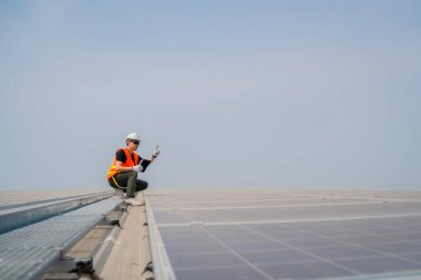 Koruyucu yelek ve beyaz şapka giyen Asyalı mühendis mavi gökyüzü arka planlı güneş paneli çatısının gücünü kontrol ediyor. Fotovoltaik teknoloji kavramı..