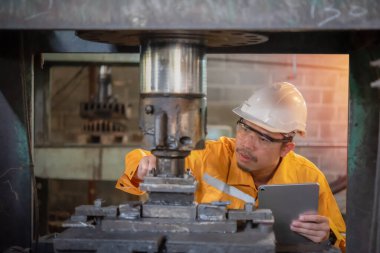 Sarı iş gözlüğü takan ve eldiven takan Asyalı mühendis tablet, sanayi ve üretim konseptini tutarken makine güvenliğini inceliyor ve denetliyor..