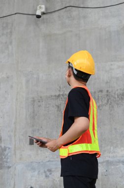 Asyalı mühendis koruyucu yelek ve şapka takıyor. İnşaat alanındaki kapalı devre kameraları kontrol etmek için tablet kullanıyor..