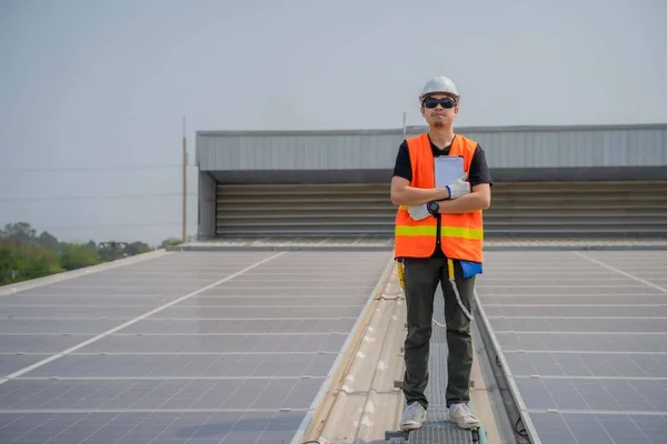 Asian engineer wearing protective vest and white hardhat standing crossed arms on solar panels roof with copy space.