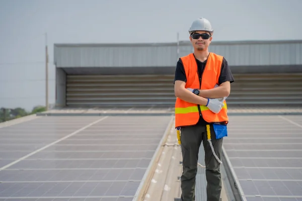 Asian engineer wearing protective vest and white hardhat standing crossed arms on solar panels roof with copy space, Photovoltaic technology concept.