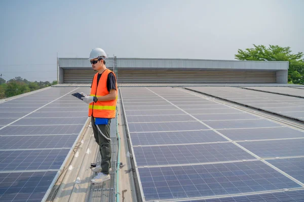 Asian engineers wearing protective vest and white hardhat standing using tablet on solar cell roof, Photovoltaic technology concept.