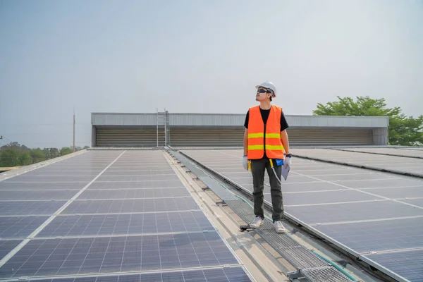 Asian engineer wearing protective vest and white hardhat standing and looking to the copy space while holding tablet on solar panels roof, Photovoltaic technology concept.