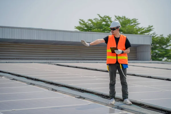 Asian engineer wearing protective vest and white hardhat standing testing heat while holding tablet on solar panels roof, Photovoltaic technology concept.
