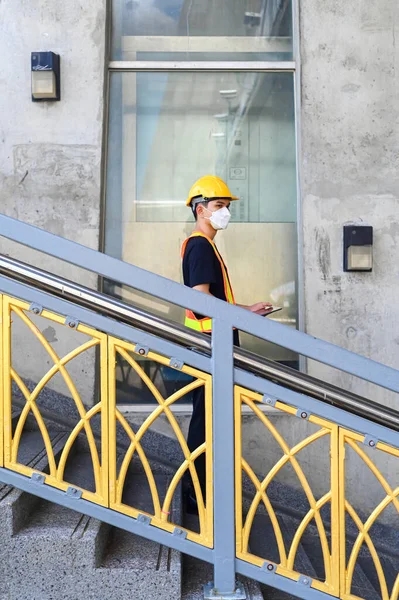 Asian engineer worker wear mask hardhat and protection vest try to check the stairs for safety.A young worker standing while holding tablet.