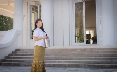 Geleneksel Taylandlı beyaz elbiseli güzel bir kadın kameraya bakıyor ve sabah gülümsemesiyle beyaz bir tapınağın önünde Pinto adında bir beslenme çantası tutuyor..