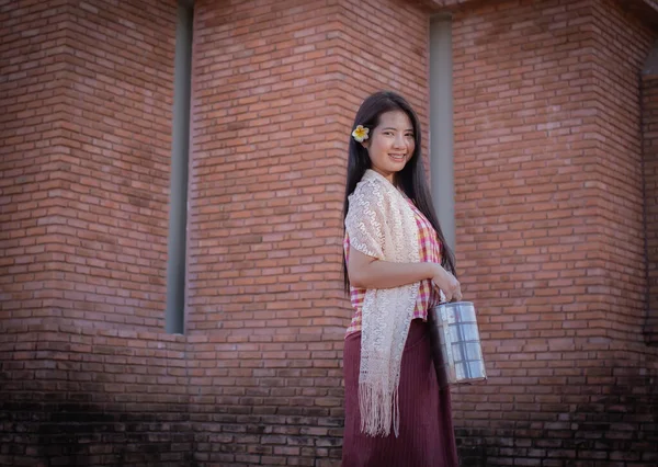 stock image Beautiful thai woman in traditional thai dress is holding her lunch box called a pinto while standing and looking at the camera in an old temple  with a smile.