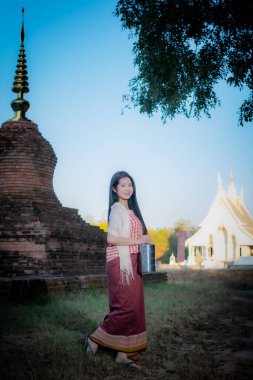 Dikey resimde geleneksel giyinmiş güzel bir Taylandlı kadın eski bir tapınakla beyaz tapınak arasında duruyor. Yüzünde bir tebessüm var.