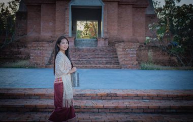 Akşam ışığında güzel bir Taylandlı kadın ayakta kameraya bakıyor ve yüzünde eski bir tapınağın önünde geleneksel bir elbise giyiyor..