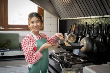 Güzel Asyalı aşçı kız önlük takıyor, elinde ahşap spatulayla tavayla mutfak arka planındaki kameraya bakıyor ve gülümsüyor..