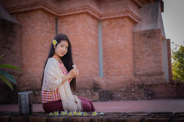 美丽的泰国人穿着传统的泰国人服装 肩上缠着白布 坐在那里 手里拿着一朵乳白色的花 脸上挂着笑容 站在一座古老的寺庙里 — 图库照片