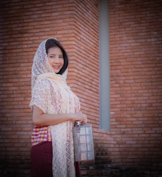 stock image Vertical picture featuring a beautiful thai girl in traditional thai dress covered with a white cloth standing and looking to the camera while holding tiffin with smile on her face in an old temple.