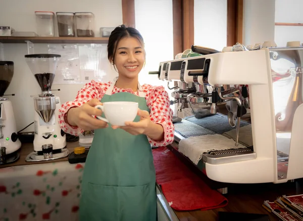 stock image Beautiful asian coffee girl wearing apron lodge a coffee cup to the camera with smile in coffee shop.