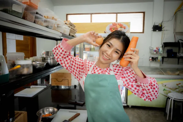 Beautiful asian female cook enjoying organic vegetables and carrots with smile in kitchen background