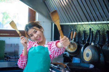 Önlük takan güzel Asyalı aşçı elinde bir spatula tutuyor ve iki eli mutfakta neşeli bir şekilde gülümsüyor..