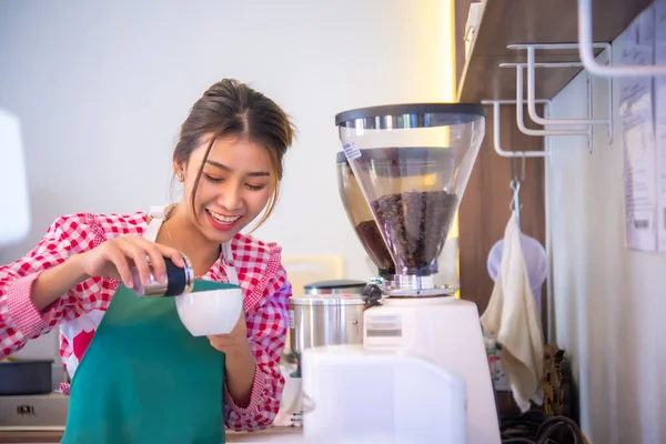 Beautiful asian coffee shop worker is looking and holding a glass and a powder jar with cheerful expression while standing next to a coffee machine, Business and finance.