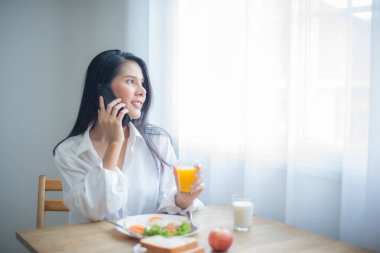 Güzel Asyalı kadın bir bardak meyve suyu içip kahvaltı ederken cep telefonuyla konuşuyordu..