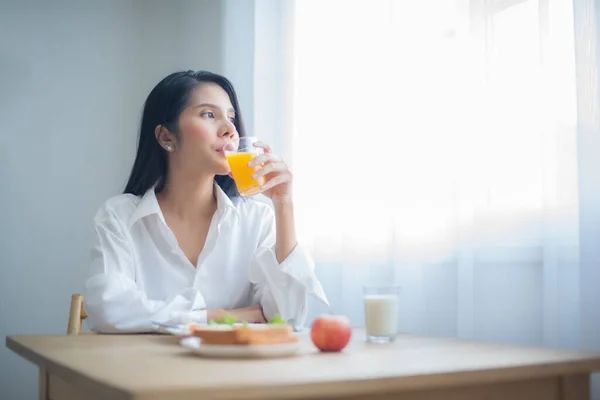 stock image Beautiful asian woman savored her glass of juice while gazing into the copy space with breakfast on the table.