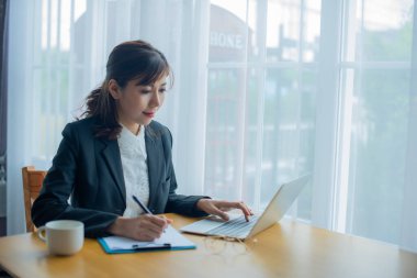 Güzel Asyalı iş kadını ofisinde bilgisayarındaki bilgilere atıfta bulunurken yüzünde gülümsemeyle bir belge yazıyor..