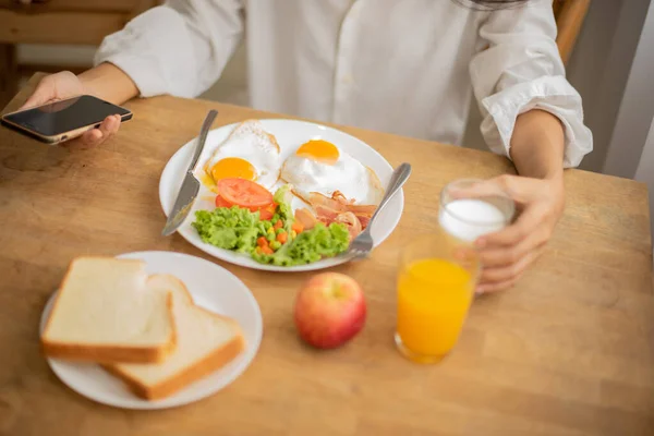 stock image Unknown person holding mobile phone and glass of milk and breakfast on table full of fresh fruit and bread.