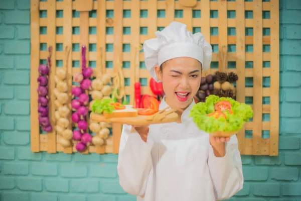 美しいアジアの女性プロのシェフがカメラを見ながら トマトパンと新鮮な緑の野菜のボウルをキッチンで楽しい表情で木製のトレイを保持します — ストック写真