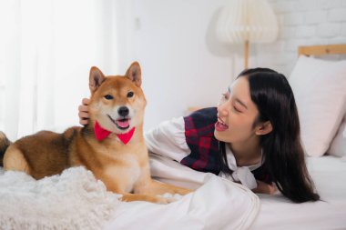 Güzel Asyalı kadın, mutlu bir ifadeyle köpeğe dokunurken beyaz yatakta uzanıyor..