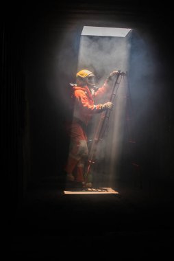 İtfaiyecinin dikey resmi, yukarıdaki delikten içeri ışığın aktığı karanlık bir odada kırmızı bir merdivene tırmanmaktır..