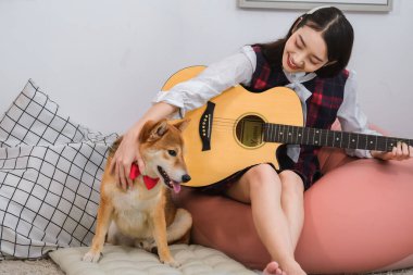 Güzel Asyalı kadın, elinde gitarıyla köpekle oturma odasında oturuyor..