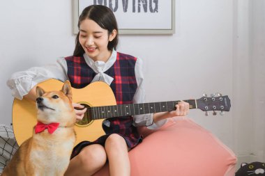 Güzel Asyalı kadın kanepede rahatça gitar çalıyor. Köpek oturma odasında otururken yüzünde gülümseme ve niyet var..