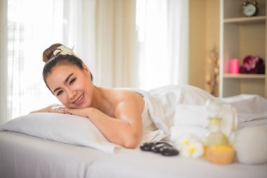 Güzel Asyalı bir kadın, spa odasındaki beyaz bir yatakta boynu eğilmiş bir şekilde uzanıyor ve yanında bir bardak kaplıca yağı var..