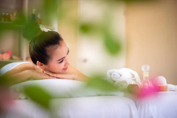 美丽的亚洲女人躺在一间舒适的水疗室的白色床上 凝视着复印的空间 眼前一片模糊的绿叶 — 图库照片