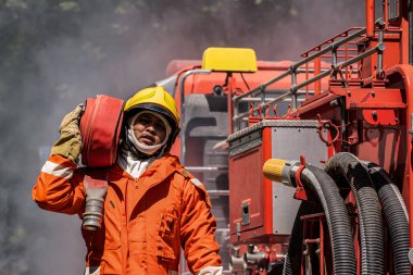 İtfaiyeci sarsılmaz bir kararlılıkla itfaiye aracının yanında dimdik durur. Güçlü tutumları, yangın hortumunu omuzlarına dayar ve harekete geçmeye hazırdır..