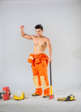 Güçlü ve kendinden emin bir pozla üstsüz bir itfaiyeci el değmemiş beyaz bir arka plana karşı duruyor elinde demir bir balta ile güç ve kararlılık yayıyor.