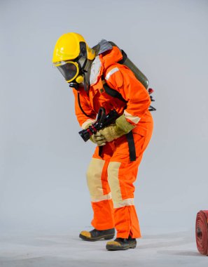 Profesyonel itfaiyeci sırtına oksijen tüpü takmış, yere şüpheyle bakıyor ve beyaz arka planda yangın hortumu tutuyor..