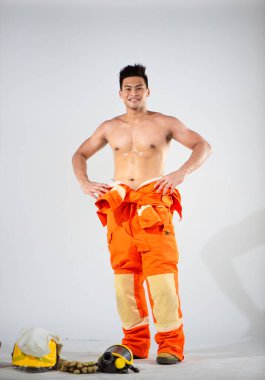 Üstsüz itfaiyeci kendine güvenen ve otoriter bir duruş sergiliyor. Temiz beyaz bir arkaplan üzerinde kollar akimbo. Güç ve dayanıklılık atmosferi yansıtıyor..