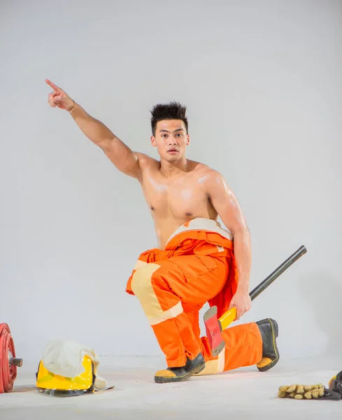 stock image Topless firefighter is sitting holding an iron axe looking at the camera and pointing a finger towards the top.