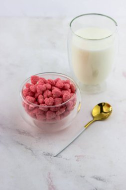 Beyaz mermer arka planda bir kase kahvaltı gevreği ve bir bardak süt. Ahududu aromalı atıştırmalıklar.