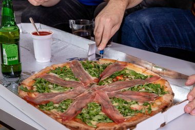 Fresh pizza with slices of ham and arugula leaves. A hand holding a knife and cutting slices from a delivery box. Home party.