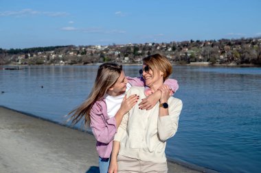 Yetişkin kızıyla gülümseyen, konuşan, eğlenen ve nehir kıyısında kumlu bir sahilde yürüyen mutlu bir anne..